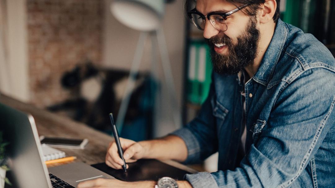 A smiling man with a beard, wearing glasses and a denim shirt, uses a graphics tablet and laptop in a modern office.