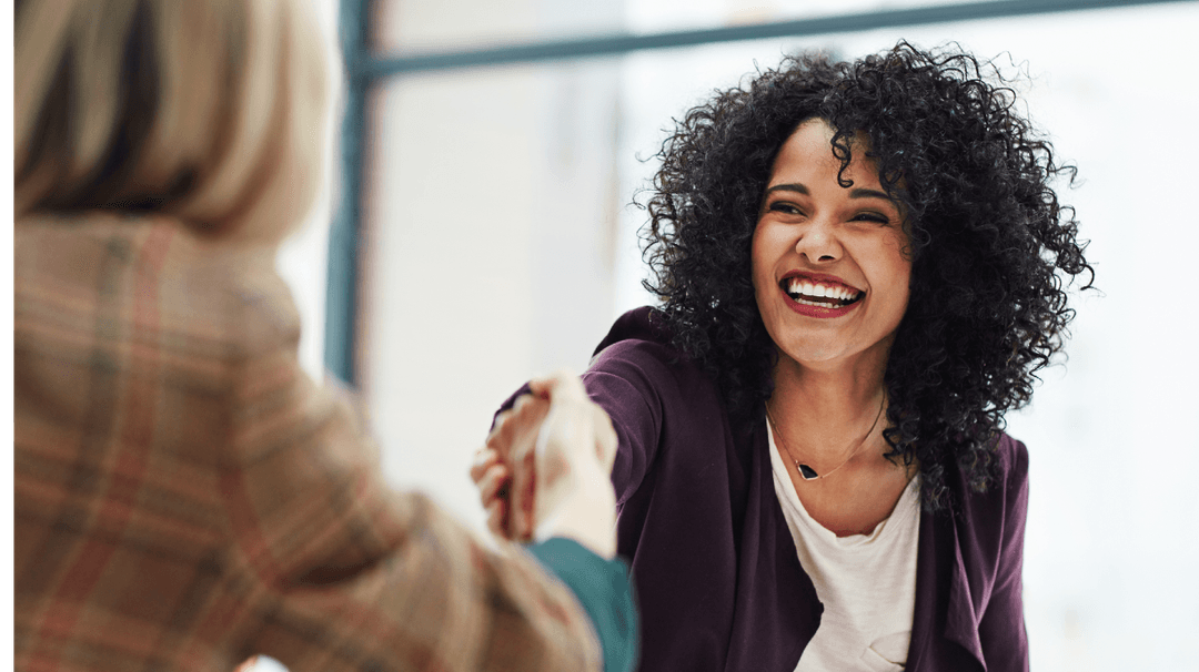 Two women in an office setting, smiling and shaking hands, one with curly hair and a purple blazer, the other partially visible.