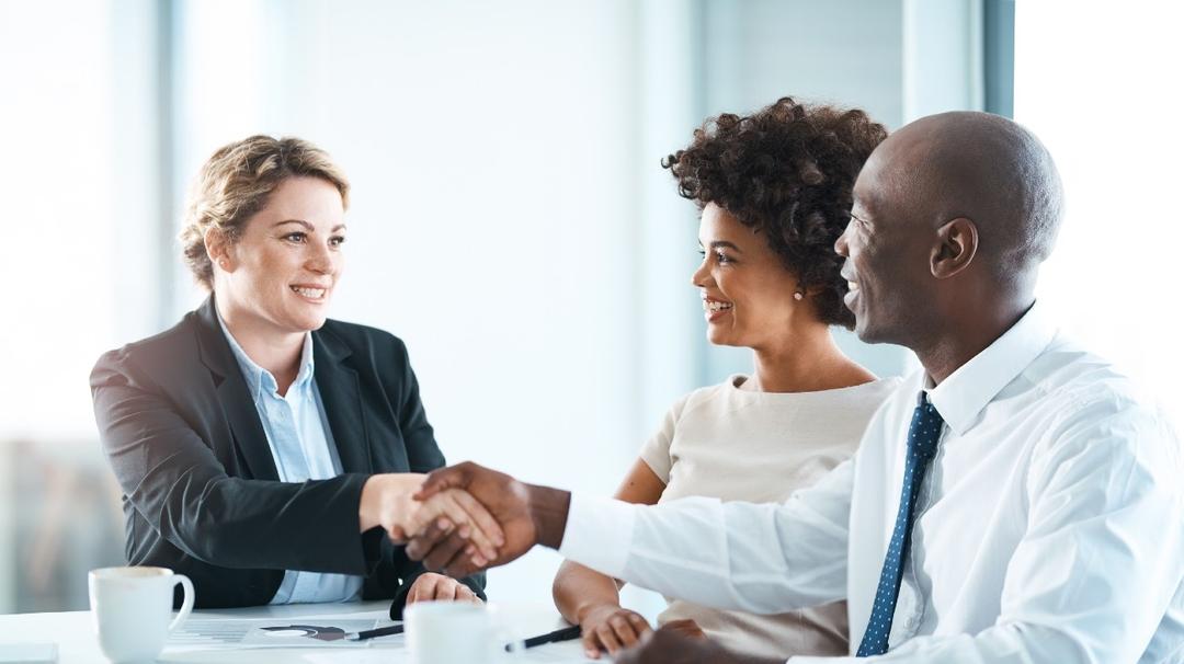 Three business professionals at a table, one caucasian woman and one african couple, smiling and shaking hands in a modern office setting.
