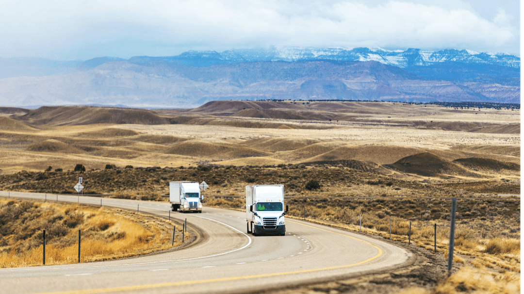 Two trucks driving on a curvy road through a desert landscape with distant snow-capped mountains under a cloudy sky.